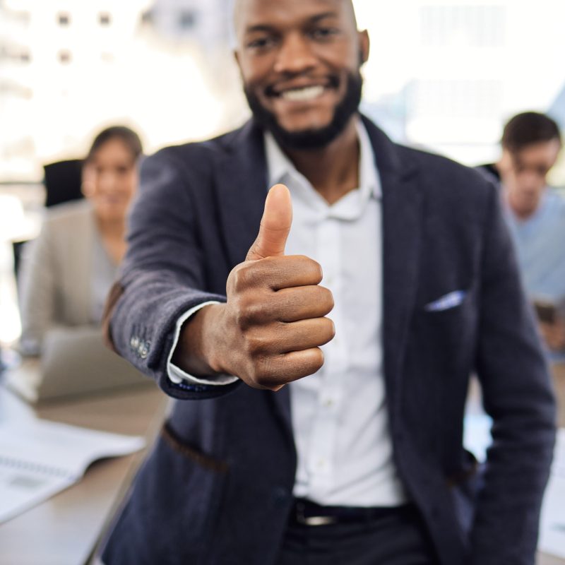 Closeup shot of a young businessman showing thumbs up in an office with his colleagues in the background.